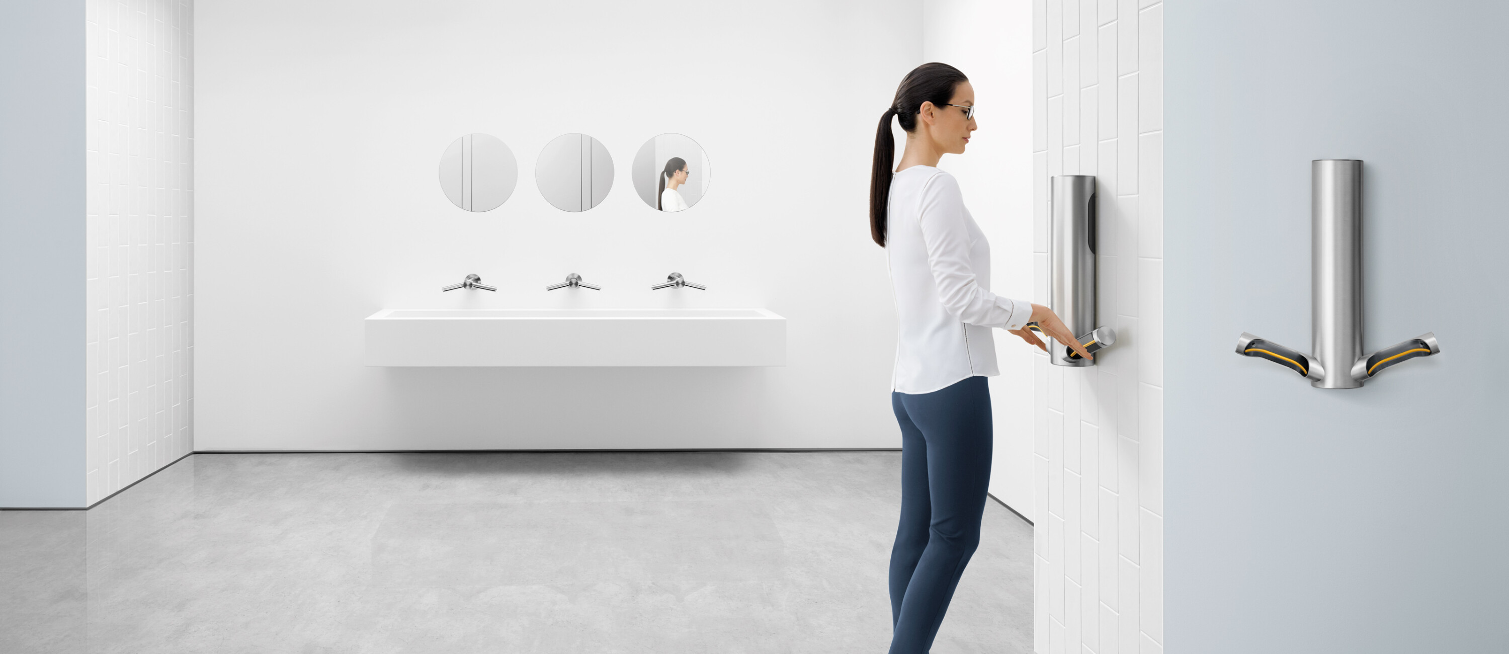 A woman with a whit shirt, blue shorts and black shoes drying her hands on a Dyson Airblade Hand Dryer Model 9kJ in a white bathroom.